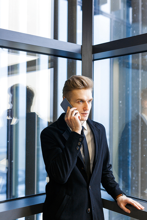 Concentrated young entrepreneur in stylish suit talking to his client on smartphone while standing by panoramic window, portrait shot