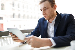 Portrait of modern businessman wearing formal suit using digital tablet at table in cafe and looking at screen