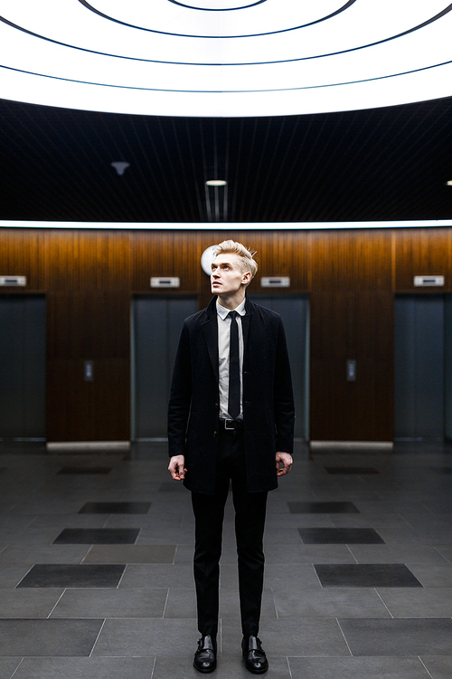 Stylish blond-haired businessman looking upwards while standing in office lobby with creative interior design, full length portrait