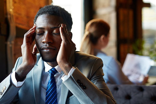 Portrait of handsome African American man wearing business suit rubbing his temples and closing eyes suffering from headache and exhaustion at table in modern restaurant