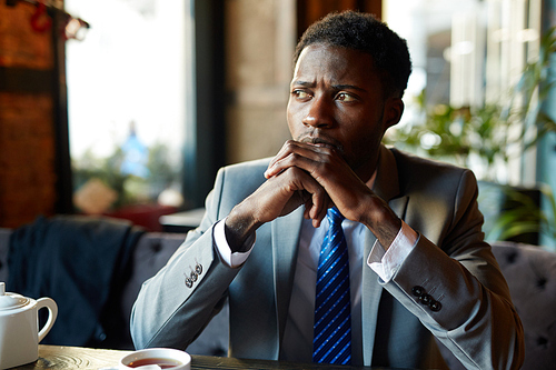 Portrait of handsome African American man wearing business suit looking away to window, pensive and worried while sitting at table in modern restaurant during coffee break