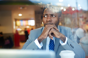 Portrait of pensive African ?American businessman looking worried and clasping hands while sitting at counter in coffee shop, shot behind glass window