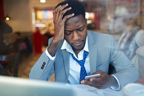 Portrait of worried African ?American businessman looking stressed and resting his head on hand while typing message in smartphone sitting at counter in coffee shop, shot behind glass window