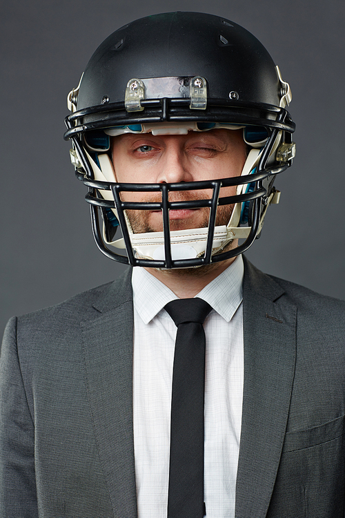 Waist up portrait of confident middle-aged businessman wearing American football helmet and suit winking at camera against grey background