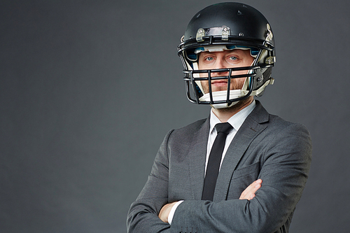 Confident businessman wearing suit and American football helmet standing cross-armed against grey background and , copy space to left
