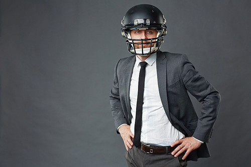 Portrait of confident businessman wearing American football helmet and suit standing with his hands on hips against grey background and  away