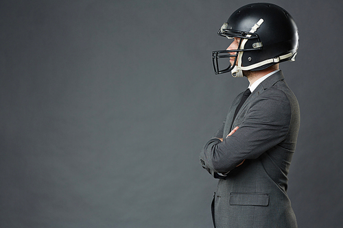 Side view of confident businessman wearing American football helmet standing cross-armed against grey background, copy space to left