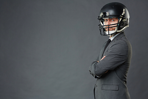 Side view of middle adult businessman wearing American football helmet standing cross-armed against grey background and , copy space to left