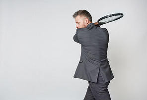 Middle aged businessman in formalwear playing tennis against grey background, copy space to the left