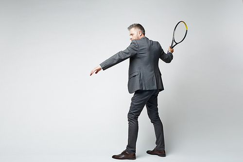 Full length portrait of middle aged businessman in suit hitting shot with tennis racquet against grey background