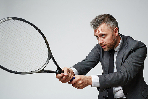 Portrait of confident middle aged businessman holding tennis racket ready to counter ball against grey background