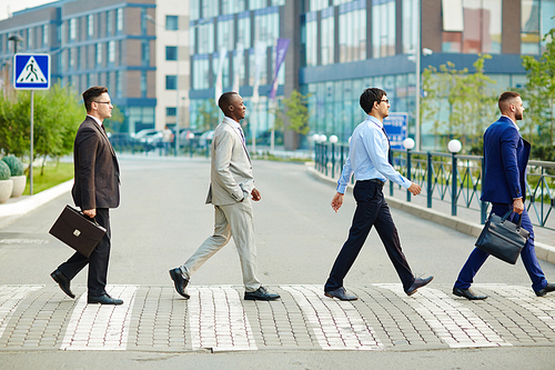 Multi-ethnic group of confident coworkers crossing road in city center, modern office buildings on background, profile view