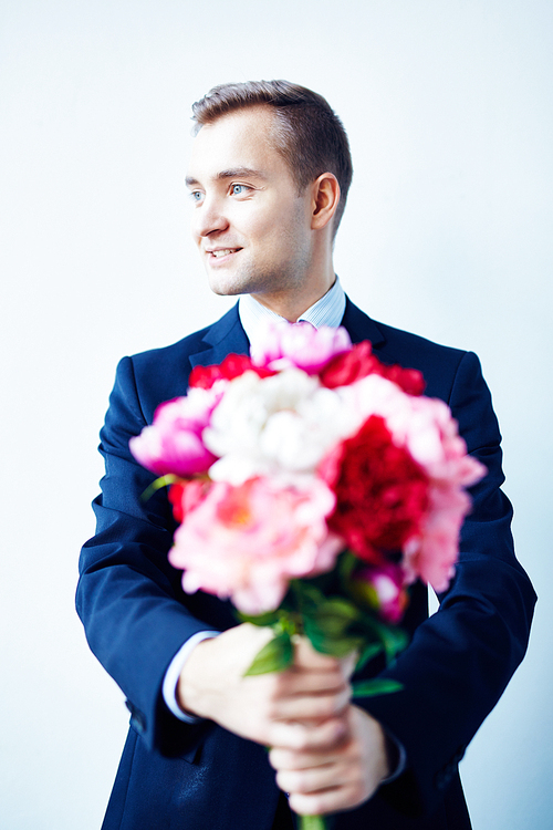 Portrait of young man in suit posing with bouquet of beautiful flowers standing against white wall