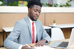 Handsome Afro-American entrepreneur wearing stylish suit sitting at desk and preparing for important negotiations with business partner, interior of open plan office on background