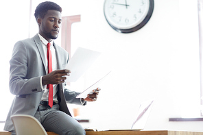 Portrait of concentrated young Afro-American businessman wearing stylish suit studying documents while working at spacious office