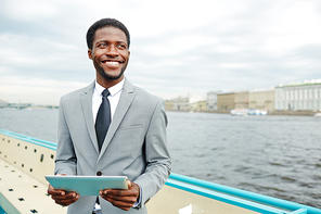 Handsome African American manager looking away with toothy smile while standing on upper deck of ship and checking business emails with help of digital tablet, waist-up portrait shot