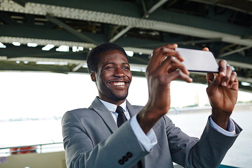 Cheerful African American entrepreneur in classical suit taking selfie on smartphone while standing on upper deck of ship, blurred background