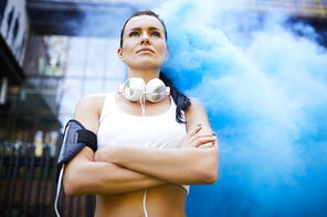 Below view of serious confident attractive sportswoman in white top wearing wired headphones on neck and looking into distance, she standing in blue smoke