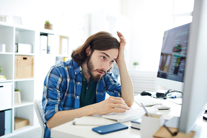 Young confused it-manager looking at one of computer monitors with tense expression