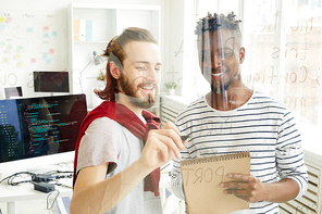 Cheerful optimistic young interracial colleagues in casual clothing writing code for computer program on glassy wall and discussing data in office