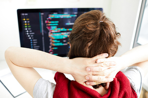 Rear view of hipster computer programmer holding hands on back of neck and relaxing while analyzing code on computer