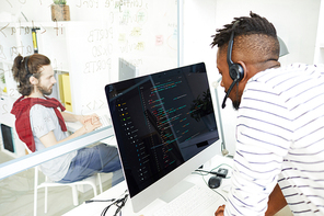 Concentrated young African-American IT support operator in headset with microphone answering question while talking to customer and using computer