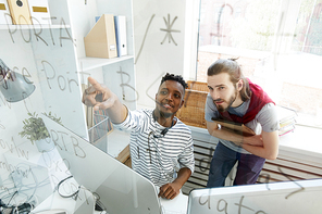 Positive confident skilled software engineers in casual clothing discussing coding algorithm: content African-American guy pointing at script on glassy board