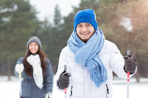 Smiling young man in blue beanie and scarf holding ski sticks and enjoying skiing in snowfall