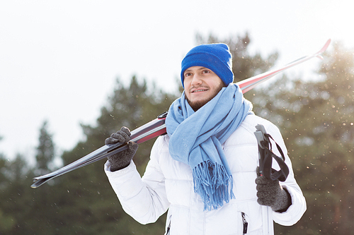 Young sportsman in white winter jacket, blue scarf and beanie hat holding pair of skis while spending time in winter park
