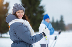 Smiling girl in grey beanie, white scarf and warm winter jacket looking at camera while skiing with her boyfriend in park or forest