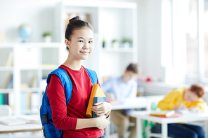 Clever and diligent Asian schoolgirl in casualwear standing in front of camera with classmates on background