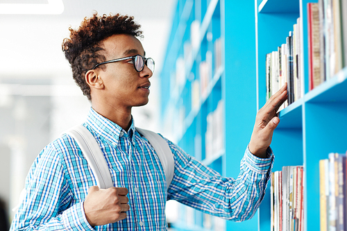 Young college student standing by bookshelf and looking for the one that is to read for next lesson