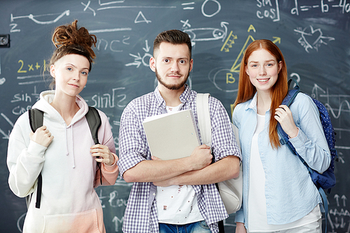 Group of three teenagers with backpacks standing against blackboard with formulae