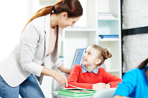 Happy young teacher talking to clever schoolgirl during individual work at lesson