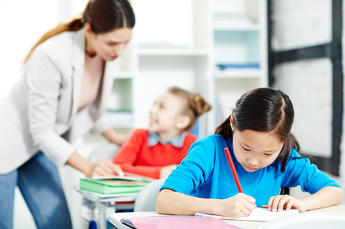 Inspired schoolgirl drawing with red crayon by desk on background of her classmate and teacher talking