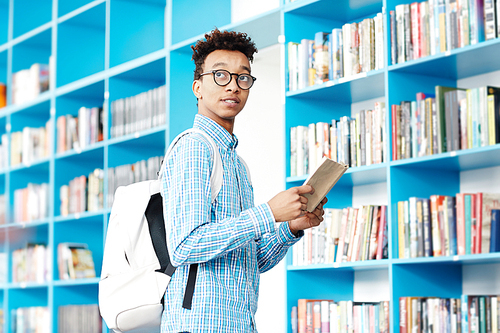 African-american student with book and backpack looking at library clock in order not to be late for next class