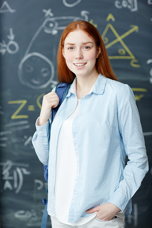 Smiling teenager with backpack standing by blackboard in classroom