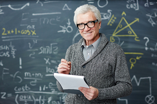 Grey-haired teacher in eyeglasses standing by blackboard with written formulae