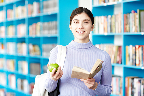Cute college girl with backpack, open book and green apple  on background of library bookshelves