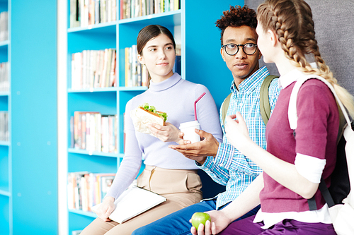 Three friendly groupmates with drinks and snack having talk at lunch break in college library