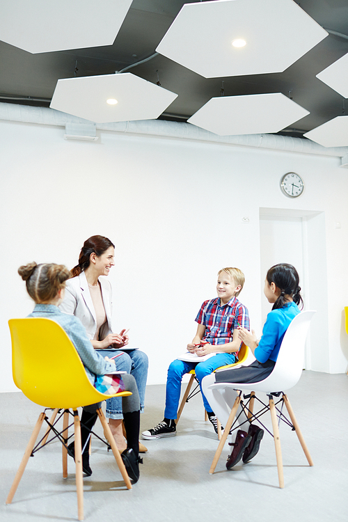 Laughing schoolkids and young teacher having talk at conversation practice lesson