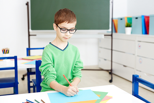 Handsome Caucasian boy in glasses sitting at classroom desk and diligently drawing on colored paper with pencil