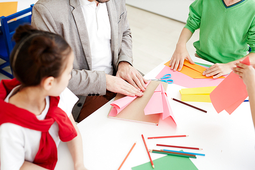 Children making paper airplanes with colored paper during art lesson while sitting at classroom desk with teacher