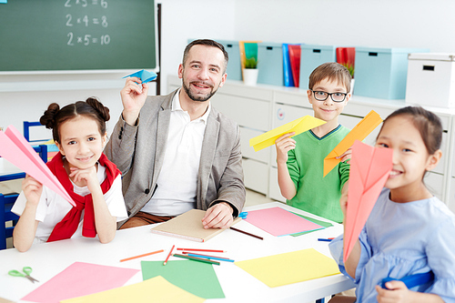 Happy pupils and their teacher with colorful paper planes sitting by table in classroom