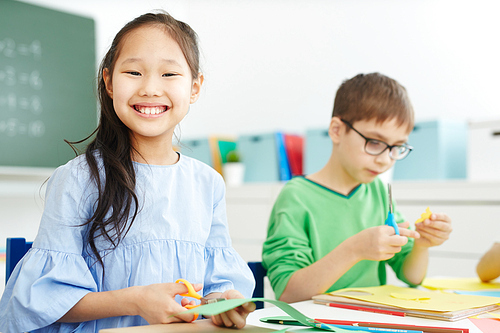 Smiling Asian schoolgirl cutting something from paper sheet at lesson of art and craft