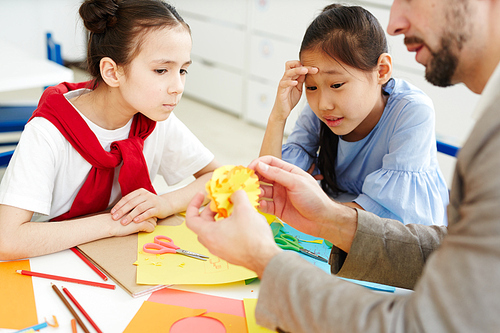 Two serious girls looking at yellow paper snowflakes in hands of their teacher