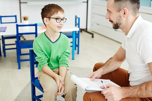 Cute schoolboy in eyeglasses answering questions of teacher at individual lesson