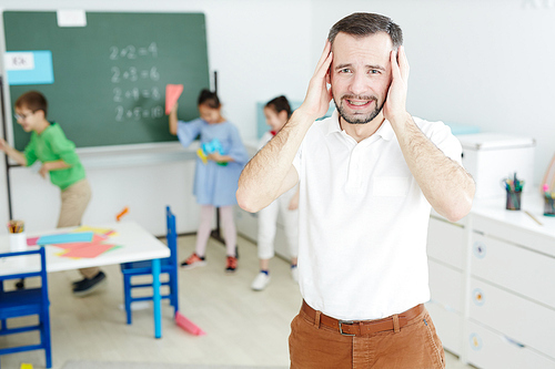 Portrait of male middle-aged art teacher standing in classroom and holding his hands on head while kids running wild with paper airplanes