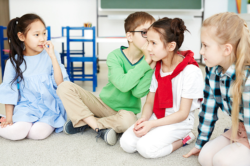 Primary school kids whispering words to each other while playing telephone game in classroom after lessons