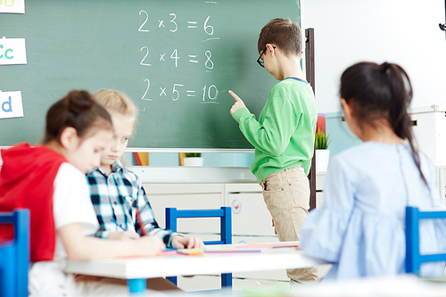 Smart little schoolboy standing by blackboard in classroom and doing multiplication sums on math class